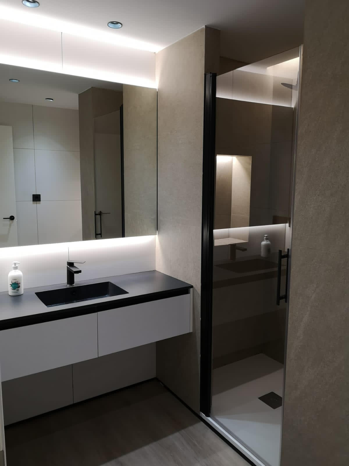 Baño con lavabo negro, espejo iluminado y ducha con mampara de cristal